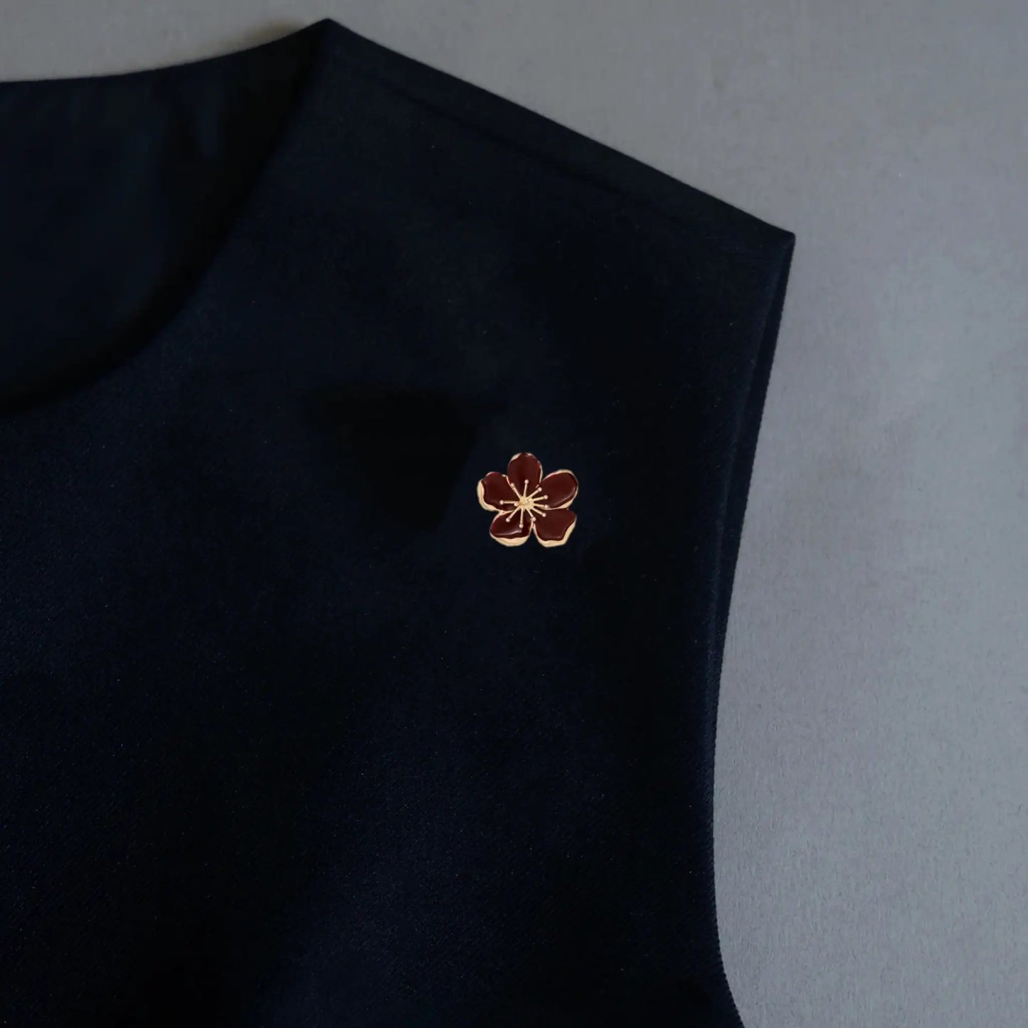 An elegant styling shot featuring a 2-in-1 burgundy wine red enamel floral accessory, worn as a sophisticated brooch pin on a black minimalist garment. The intricate gold metallic stamen adds a refined, vintage quiet luxury touch to the dark outfit.