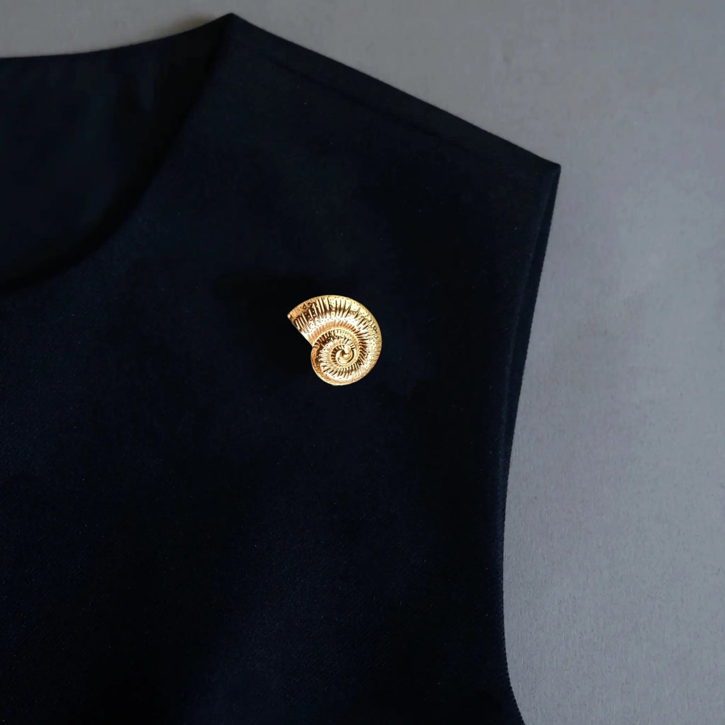 An elegant styling shot featuring a 2-in-1 textured gold nautilus shell accessory worn as a chic brooch pin on a black sleeveless garment. This realistically proportioned ocean-inspired piece adds a sophisticated statement and a touch of quiet luxury to any dark outfit.