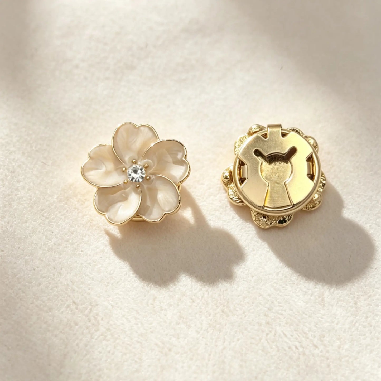 Close-up of a creamy white floral button cover showcasing a subtle marbled enamel texture. The right side reveals the closed gold back clasp, perfectly styled on textured fabric under warm sunlight.
