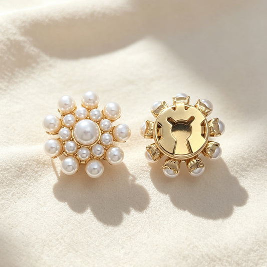 A pair of light luxury pearl cluster floral button covers resting on soft cream fabric under warm sunlight. The right piece highlights the secure closed gold backing, demonstrating its seamless use as a sophisticated faux cufflink accessory.