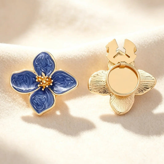 A close-up of a lapis blue marbled enamel four-petal floral button cover with a detailed textured gold stamen. Displayed on a soft cream fabric, the right piece shows the open gold clasp mechanism, demonstrating how this faux cufflink easily slides over standard buttons for a quiet luxury upgrade.