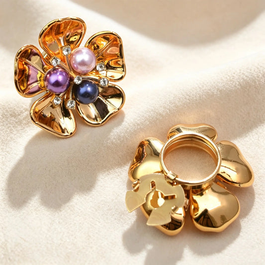 Luxurious polished gold floral button cover featuring three multi-colored pearls in pink, purple, and dark tones. The back displays a gold open clip mechanism for effortless and eye-catching styling.