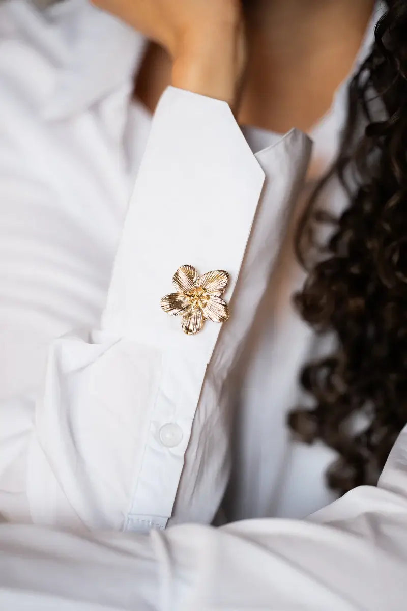 An elegant close-up of a crisp white shirt with the cuff folded up, accented by a single textured gold five-petal floral button cover. This image showcases how to effortlessly elevate a basic white button-down into a stunning piece of minimalist fashion.