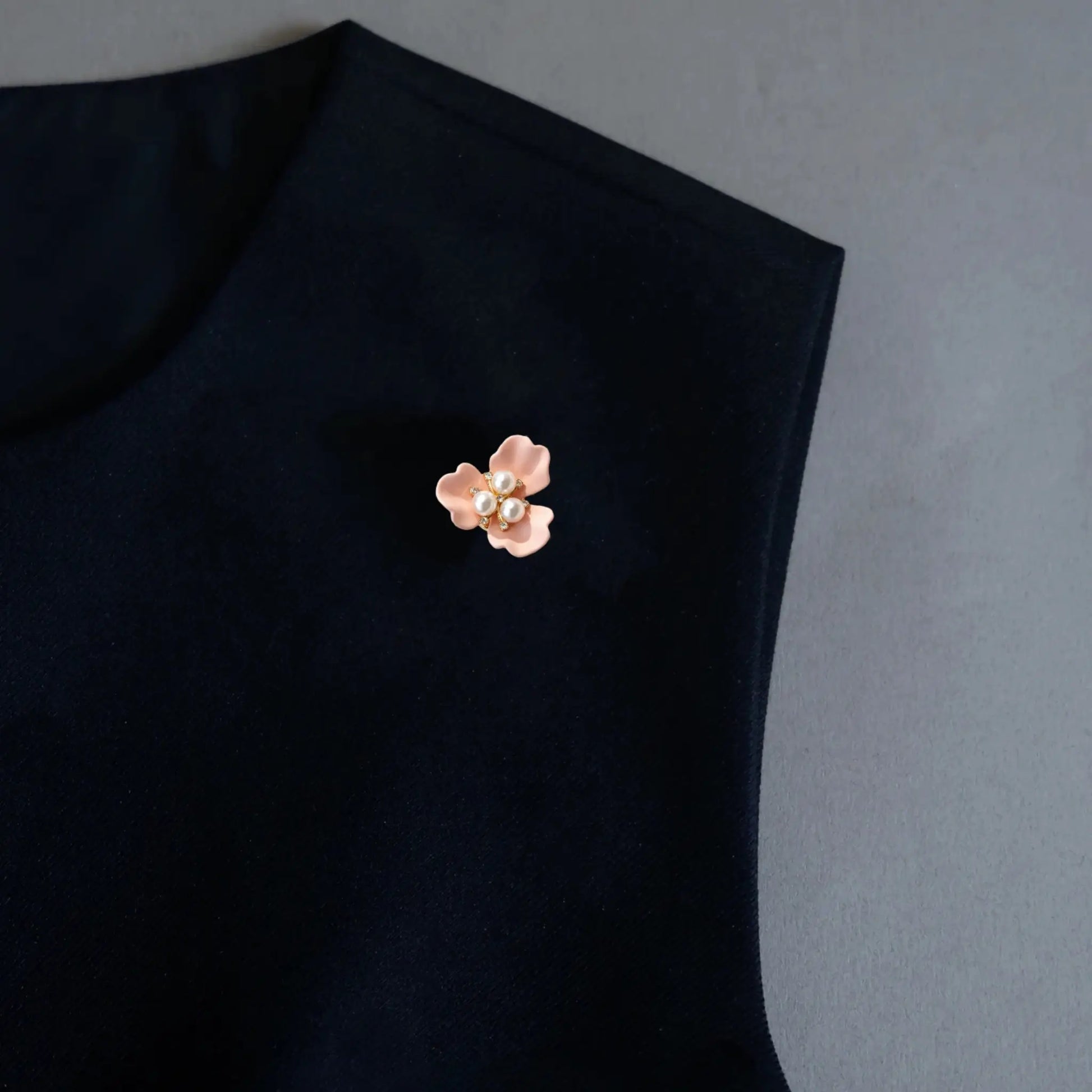 A refined minimalist styling shot displaying a versatile 2-in-1 blush pink enamel floral accessory worn as a lapel pin on a black garment. Accented with a three-pearl center, this delicately scaled piece offers a soft pop of color and an understated elegant statement for modern everyday wear.