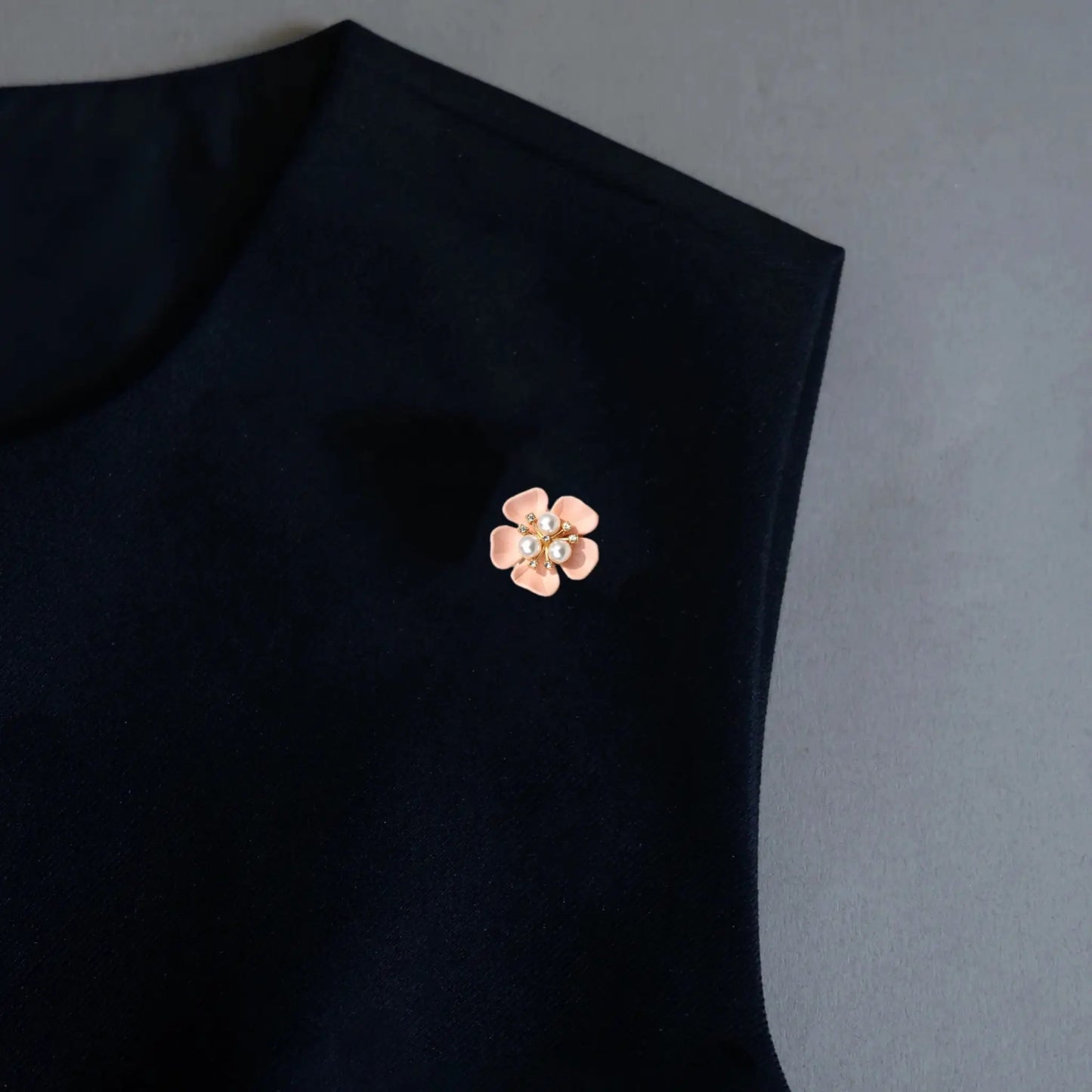 An elegant styling image featuring a versatile 2-in-1 dusty rose enamel floral accessory worn as a delicate brooch pin on a black minimalist garment. Accented with a pearl cluster center, this piece adds a soft, quiet luxury touch to any dark outfit.