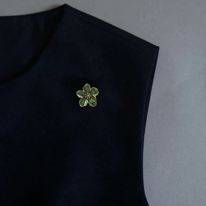 An elegant styling image displaying a versatile 2-in-1 marbled green enamel floral accessory worn as a chic brooch on a dark minimalist garment. Featuring a textured stamen and vintage color, this piece adds a unique, quiet luxury statement to modern outfits.