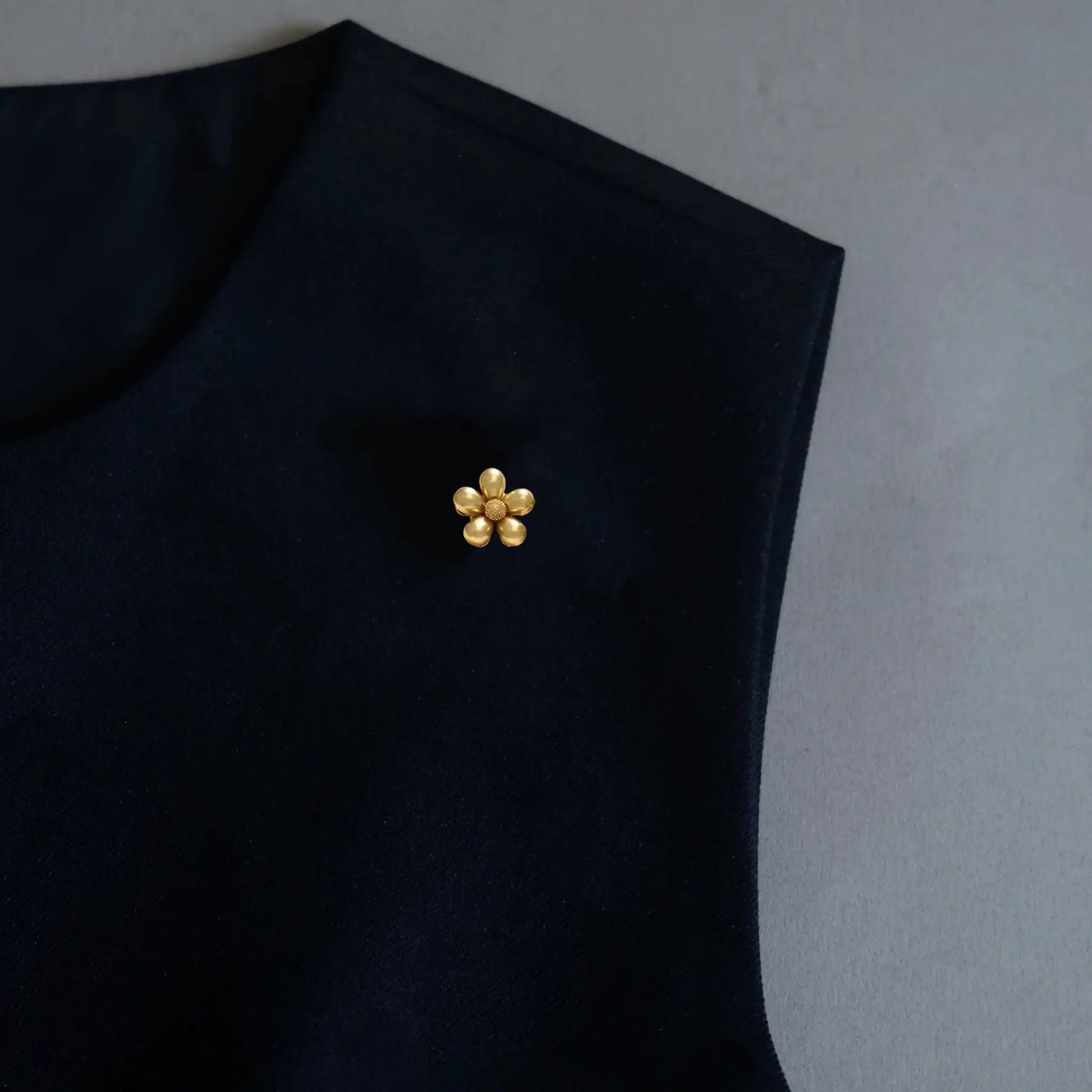 A refined styling image displaying a versatile 2-in-1 matte gold floral accessory elegantly utilized as a chic brooch on a dark top. The petite five-petal design and frosted gold finish perfectly embody the old money aesthetic for a sophisticated, minimalist wardrobe.
