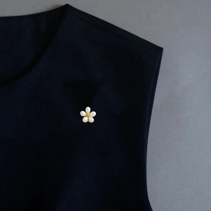 An elegant minimalist styling shot featuring a 2-in-1 petite matte pearl floral accessory, worn as a subtle lapel pin on a black garment. With its frosted pearl finish and gold stamen center, it adds a touch of understated elegance and quiet luxury to any everyday outfit.