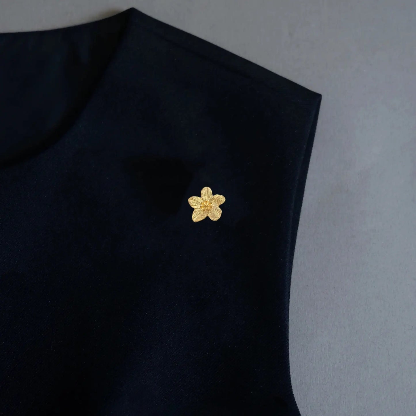 A refined minimalist styling shot featuring a 2-in-1 petite textured gold floral accessory, elegantly worn as a delicate brooch pin on a black garment. This finely crafted, light luxury piece perfectly embodies the quiet luxury aesthetic, offering a subtle yet sophisticated statement for modern wardrobes.