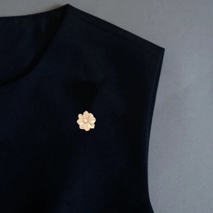 A refined styling photo demonstrating a versatile 2-in-1 textured gold floral accessory, elegantly utilized as a chic brooch on a dark top. Featuring delicate metallic brushed details and a single pearl, it provides a timeless, quiet luxury statement for evening wear.