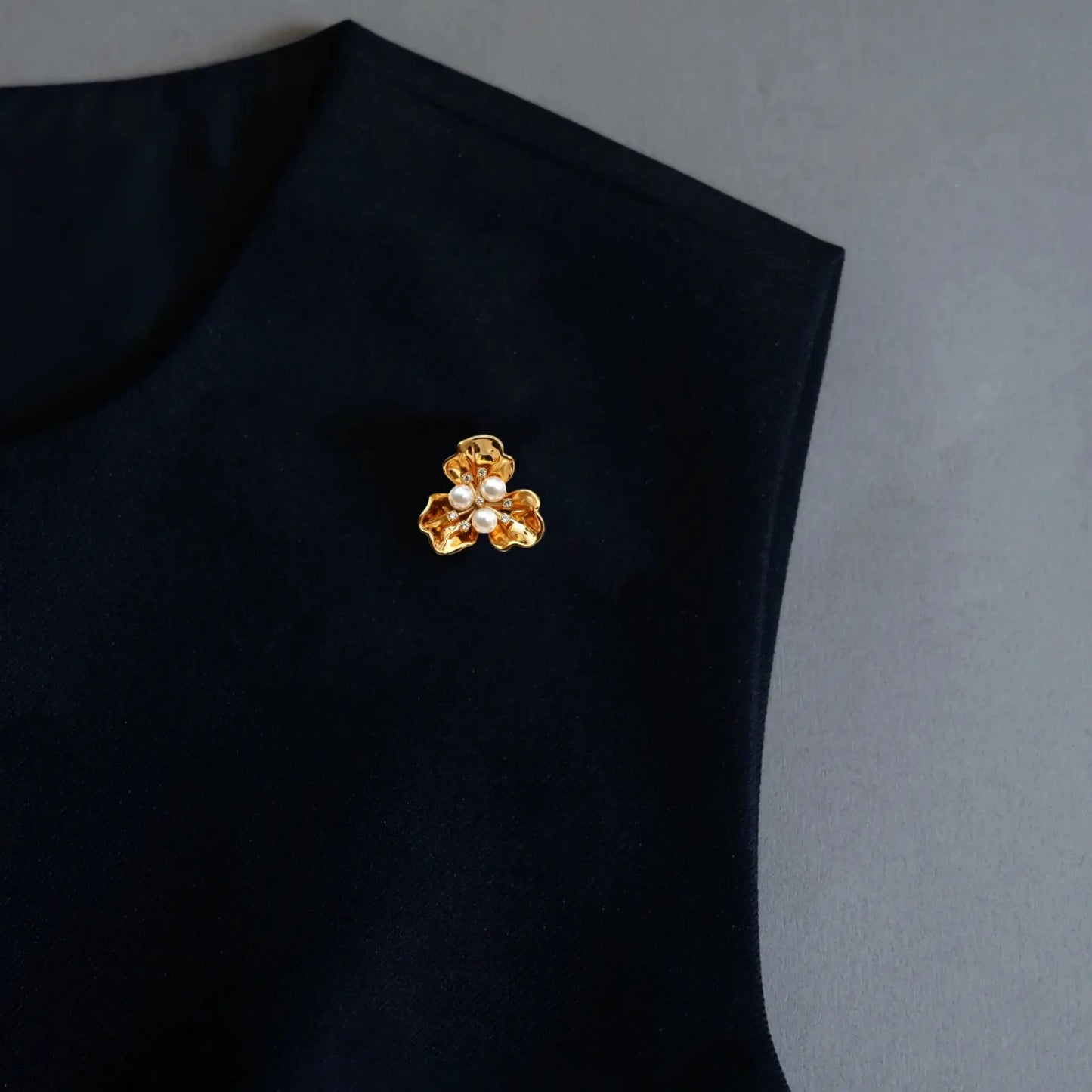A sophisticated styling image showcasing a 2-in-1 textured gold floral accessory utilized as a lapel pin on dark clothing. Featuring three central pearls and intricate metallic details, this perfectly proportioned piece embodies the old money aesthetic for modern fashion.