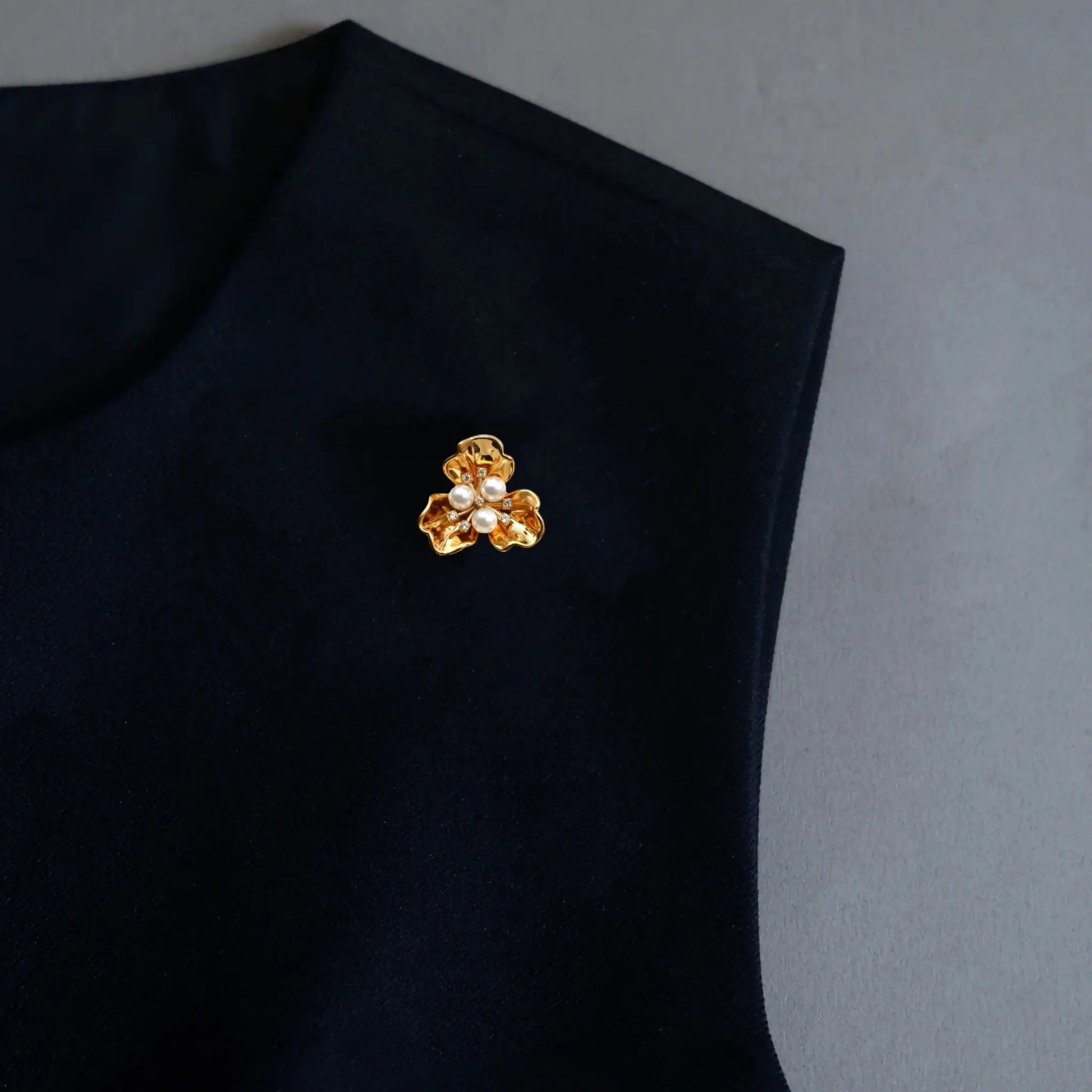 A sophisticated styling image showcasing a 2-in-1 textured gold floral accessory utilized as a lapel pin on dark clothing. Featuring three central pearls and intricate metallic details, this perfectly proportioned piece embodies the old money aesthetic for modern fashion.