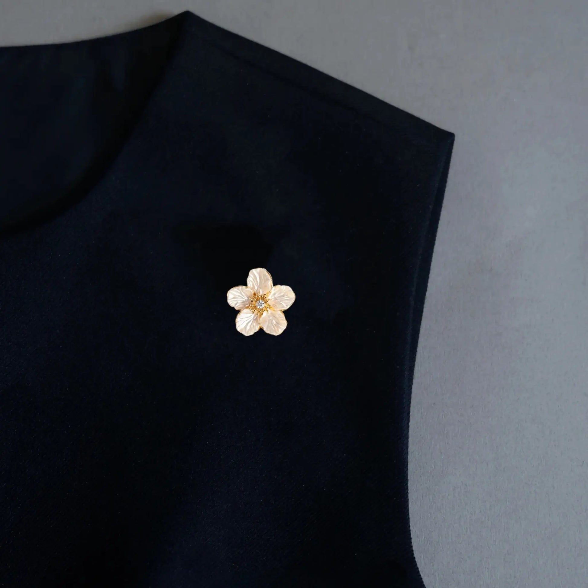 An elegant minimalist styling shot featuring a 2-in-1 textured white floral accessory worn as a delicate lapel pin on a black garment. With its natural petal texture and gold center, this versatile piece adds a refined, light luxury touch to modern everyday wardrobes.