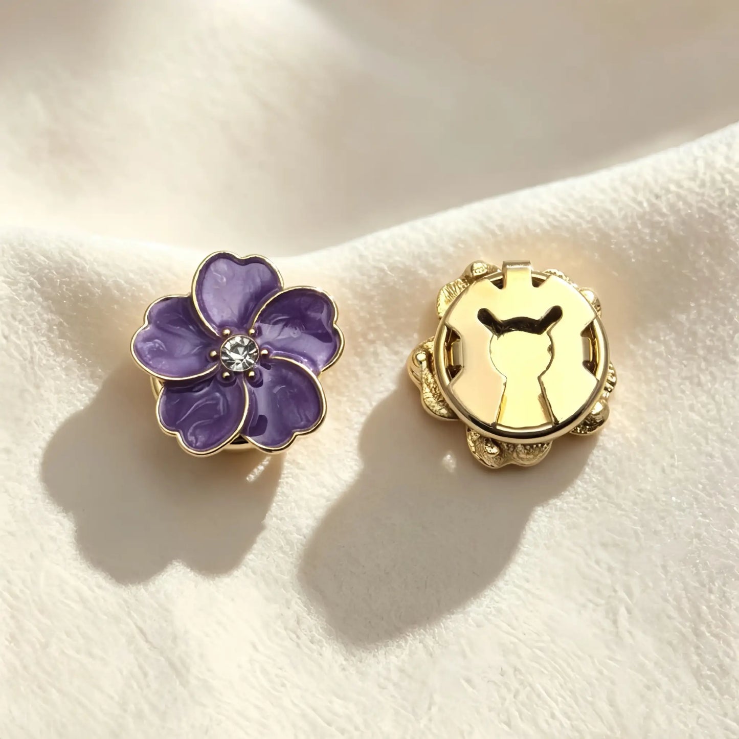 Detailed view of an amethyst purple floral button cover with a glossy enamel finish. Resting on soft cream fabric, the image highlights the secure closed gold backing for a seamless shirt upgrade.