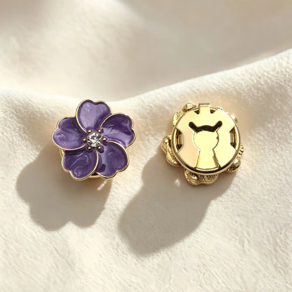 Detailed view of an amethyst purple floral button cover with a glossy enamel finish. Resting on soft cream fabric, the image highlights the secure closed gold backing for a seamless shirt upgrade.