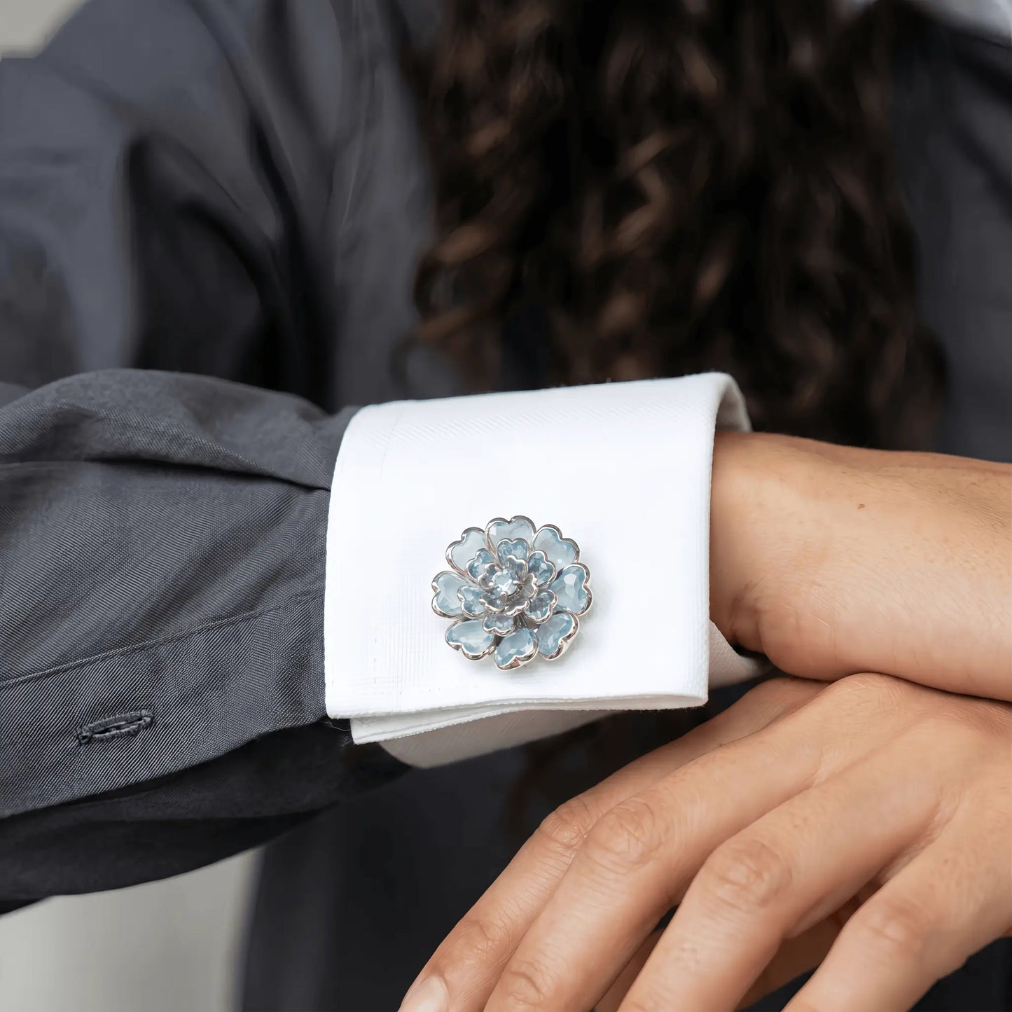A sophisticated lifestyle shot showcasing a layered aquamarine blue floral button cover worn on the crisp white contrast cuff of a charcoal grey shirt. An elegant and professional faux cufflink solution, perfect for adding a refined touch of quiet luxury to modern workwear.