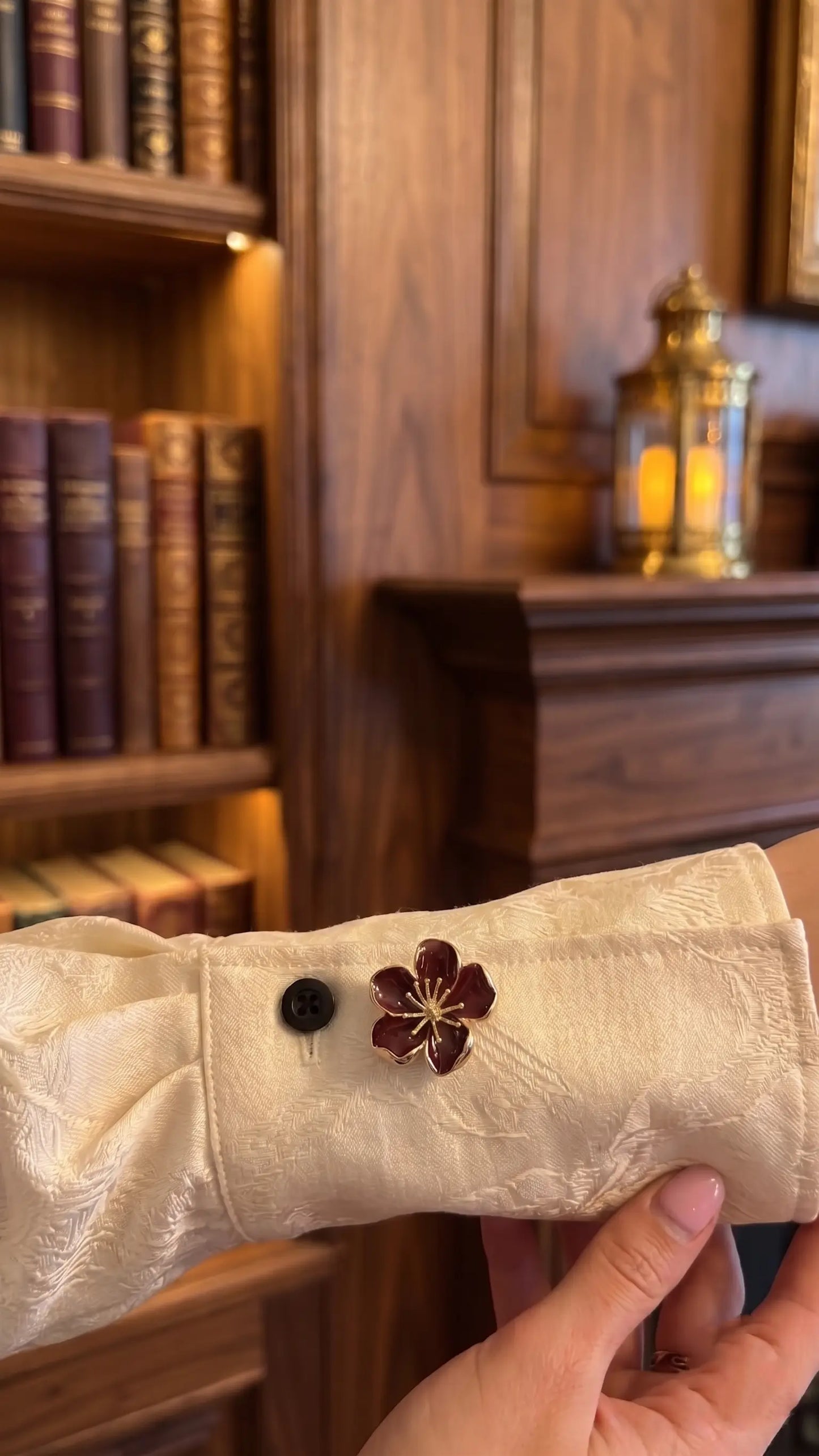 A close-up lifestyle image featuring a burgundy wine red enamel floral button cover with a gold stamen, worn on a textured cream blouse. Set against a warm, vintage library background with classic books, it perfectly captures the dark academia and old money aesthetic.