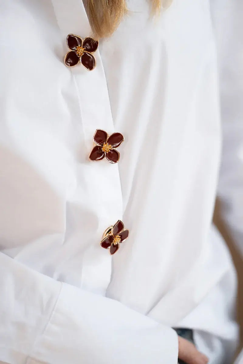 A creative styling lifestyle shot of a woman wearing a classic white shirt, uniquely utilizing three burgundy enamel four-petal floral button covers along the front shirt placket. This clever no-sew clothing hack demonstrates the incredible versatility of our button covers beyond just the cuffs.