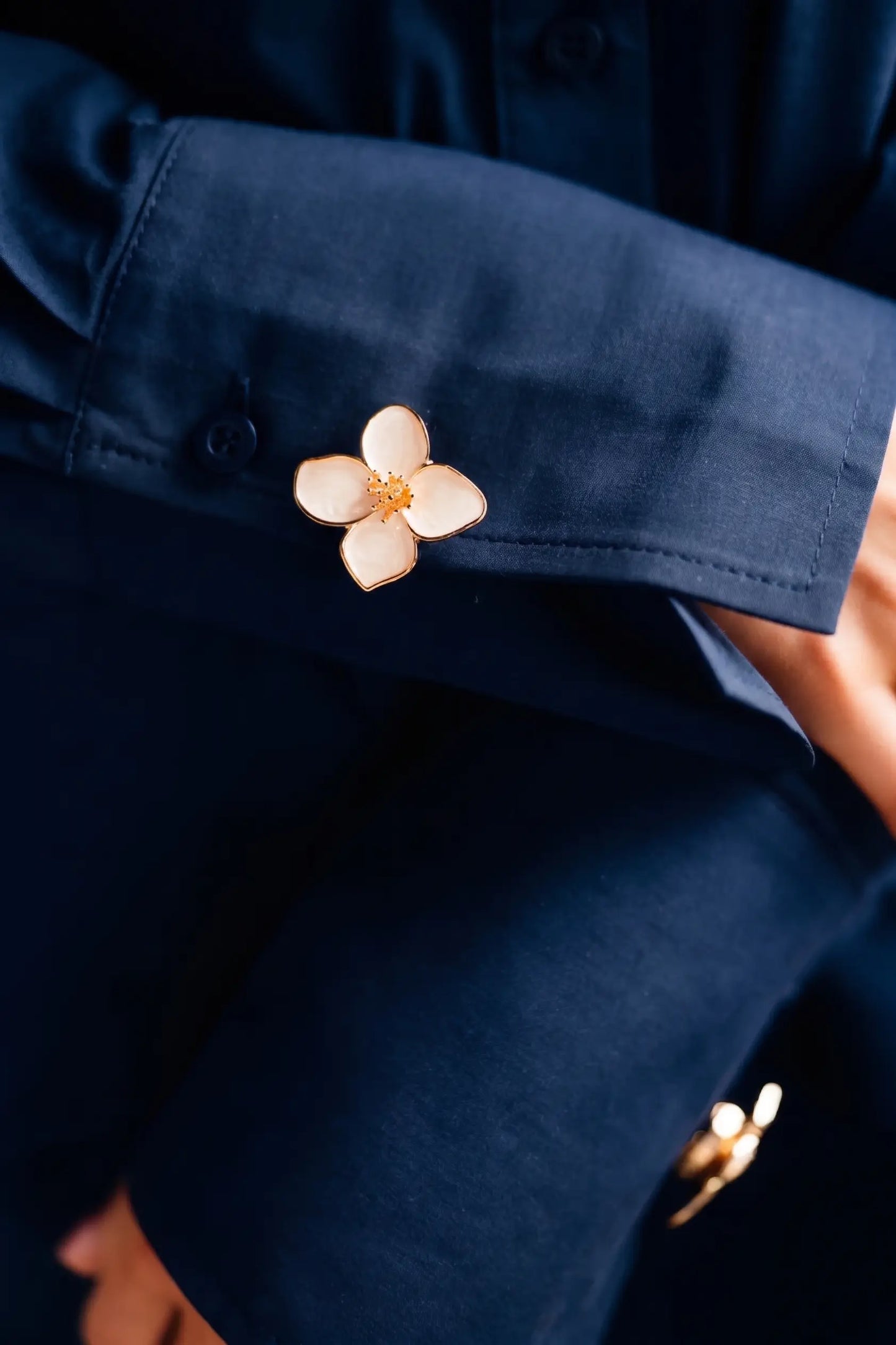 A refined styling detail shot showing a cream white enamel four-petal floral button cover with a gold stamen, smoothly attached to a navy blue shirt cuff. It demonstrates a perfectly proportioned, classic preppy look suitable for any sophisticated daily wardrobe.
