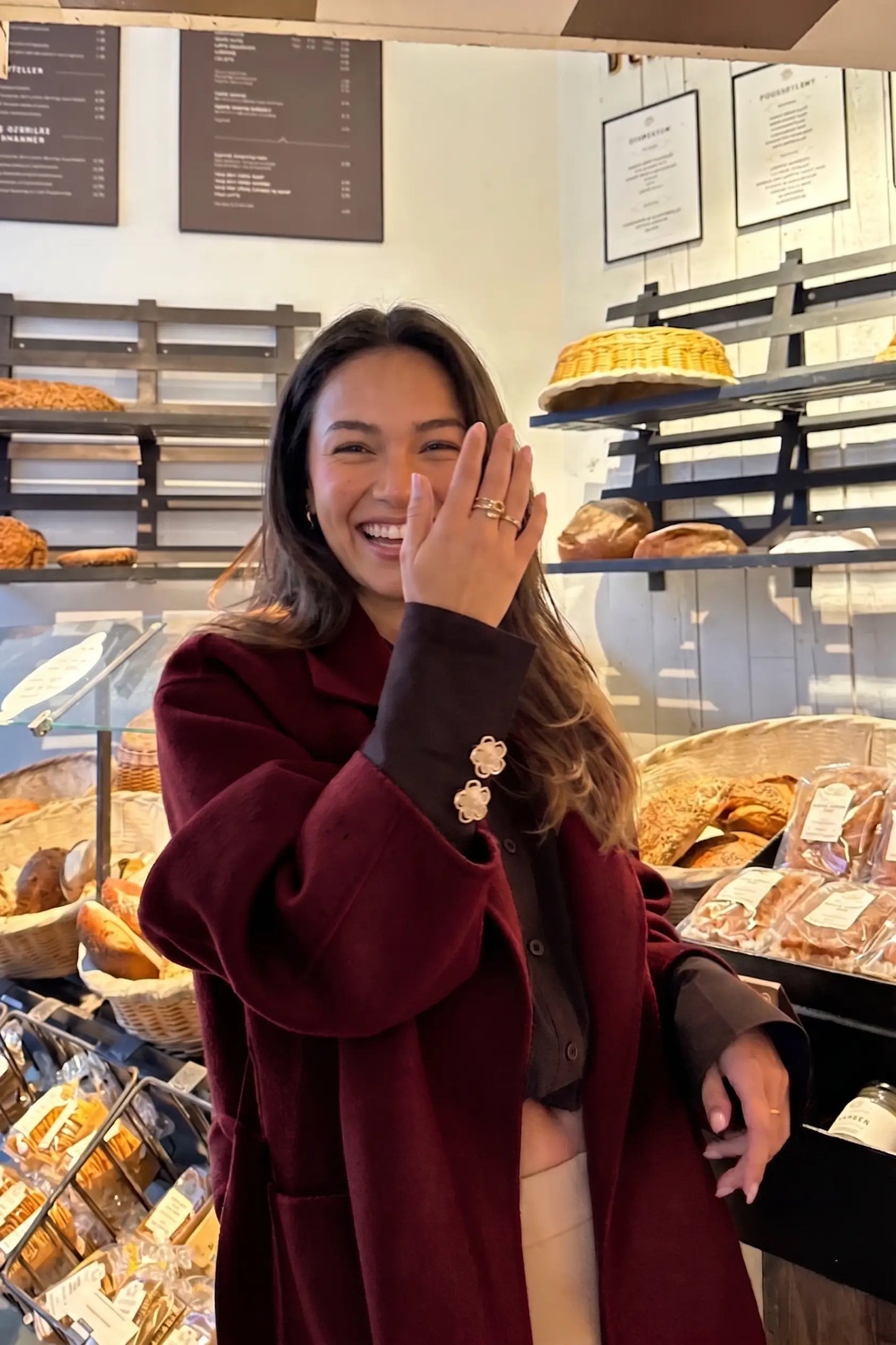 candid lifestyle shot of a laughing woman in a bakery wearing a burgundy coat with black cuffs. The cuffs are playfully styled with two cream white floral button covers, demonstrating how to easily add a touch of Parisian chic and everyday luxury to a casual winter outfit.