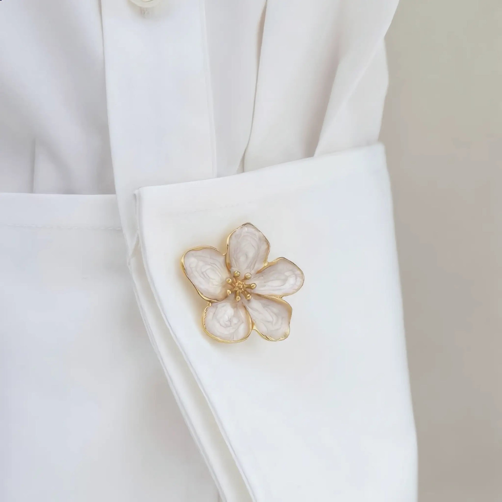 A close-up, authentic lifestyle shot of a white shirt sleeve effortlessly upgraded with a creamy white swirl enamel floral button cover. Featuring a textured gold center and a realistically small, refined proportion, this faux cufflink adds a touch of everyday chic to standard button-down shirts.