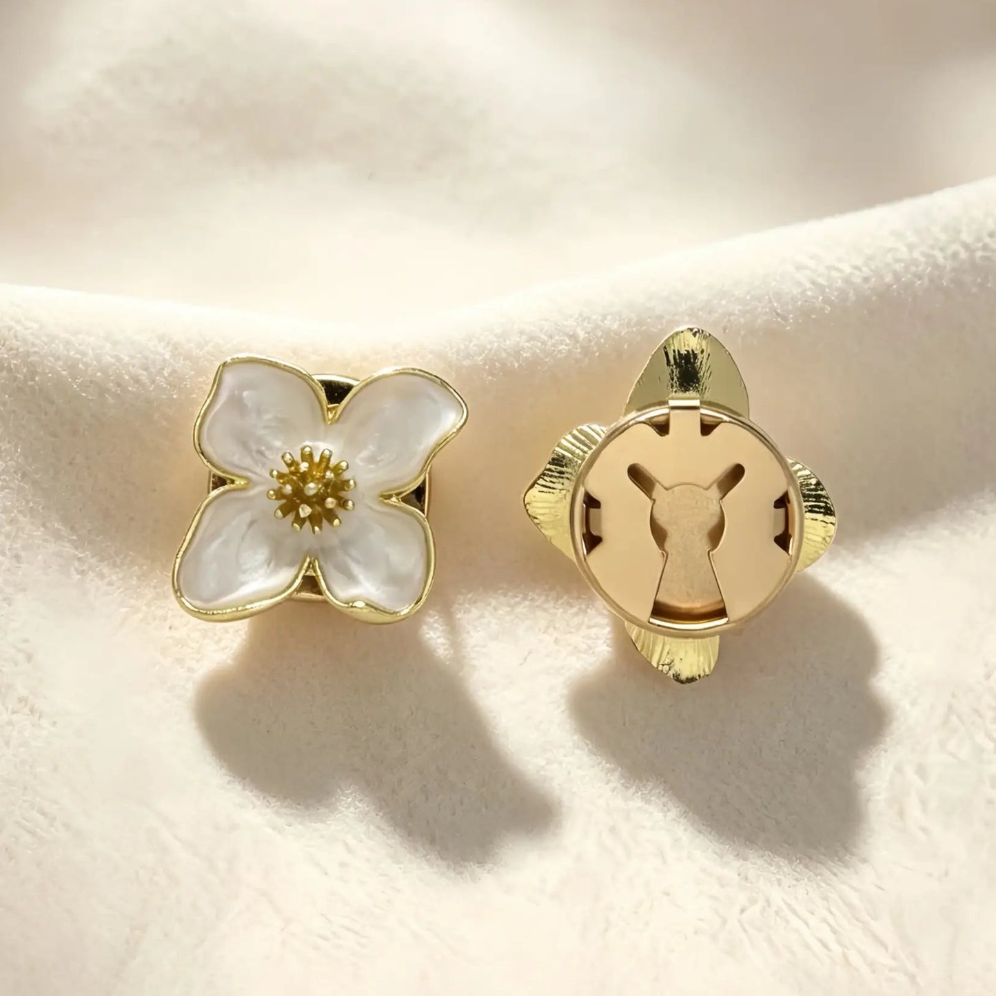A pair of creamy white enamel four-petal button covers resting gracefully on textured fabric. The image shows the aesthetic front design and the secure closed gold back, ideal for transforming standard buttons into French-style cuffs.
