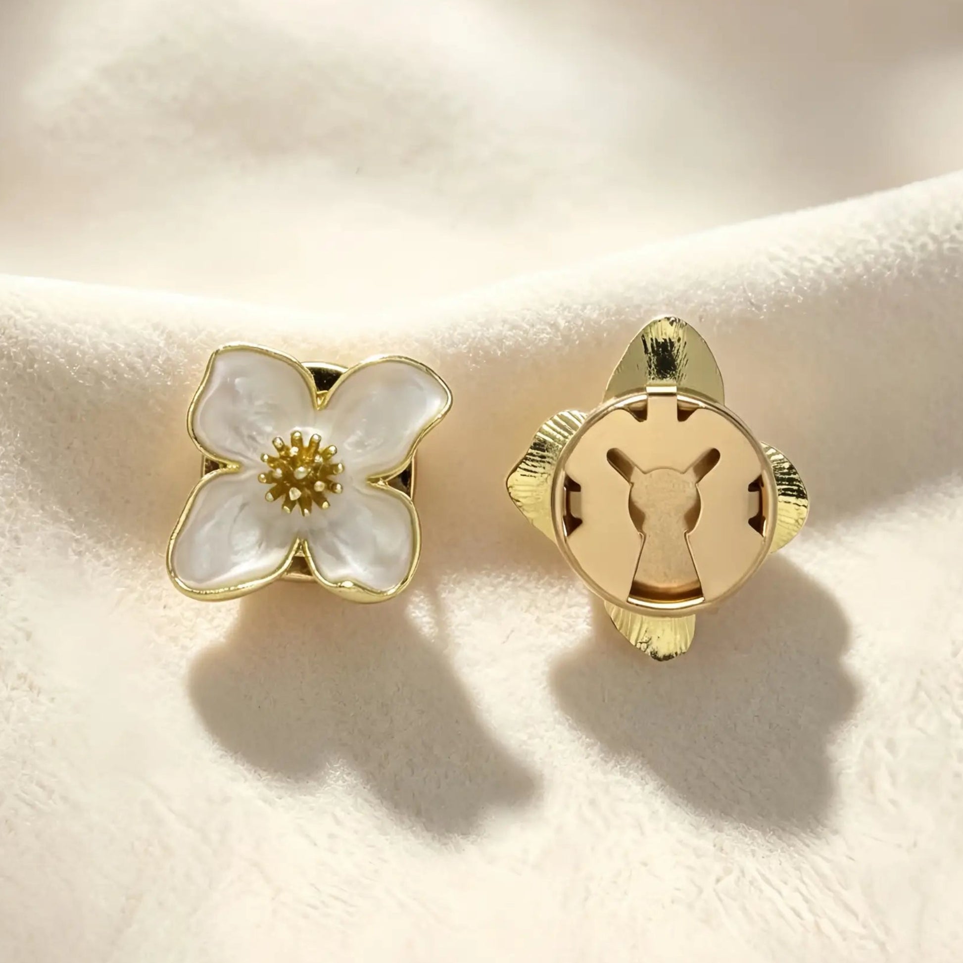 A pair of creamy white enamel four-petal button covers resting gracefully on textured fabric. The image shows the aesthetic front design and the secure closed gold back, ideal for transforming standard buttons into French-style cuffs.