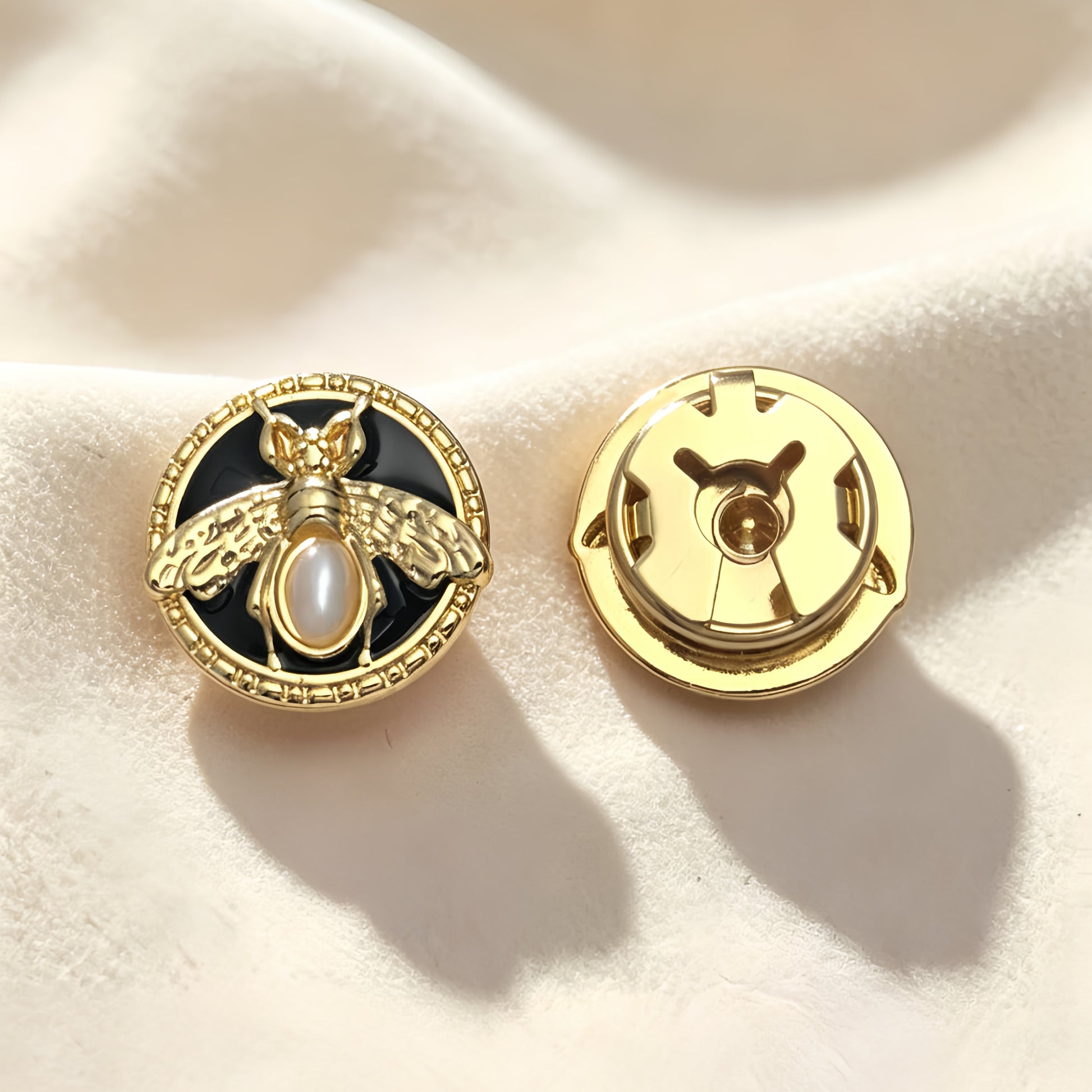 Detailed view of a light luxury black and gold bee button cover resting on soft cream fabric under warm sunlight. The right piece highlights the secure closed gold backing, demonstrating its flawless use as an elegant heritage-style cuff accessory.