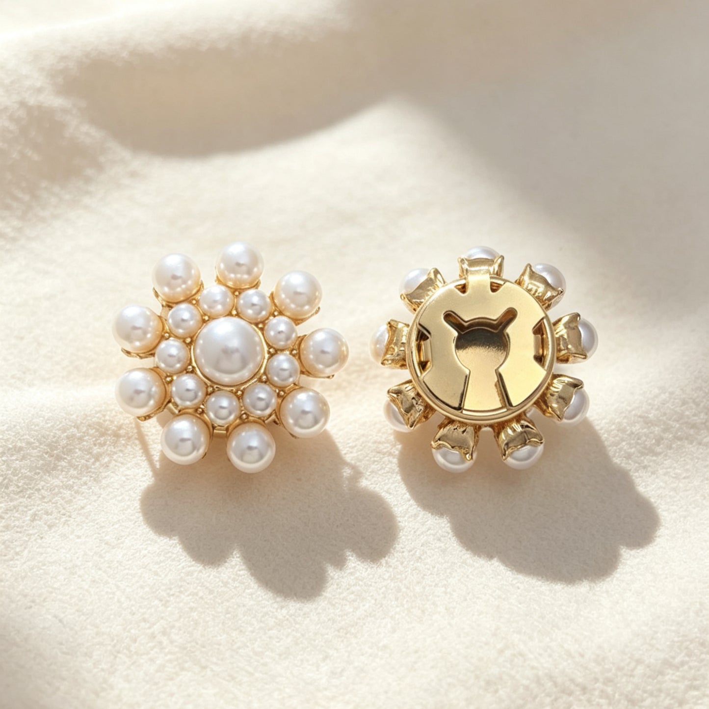 A pair of light luxury pearl cluster floral button covers resting on soft cream fabric under warm sunlight. The right piece highlights the secure closed gold backing, demonstrating its seamless use as a sophisticated faux cufflink accessory.