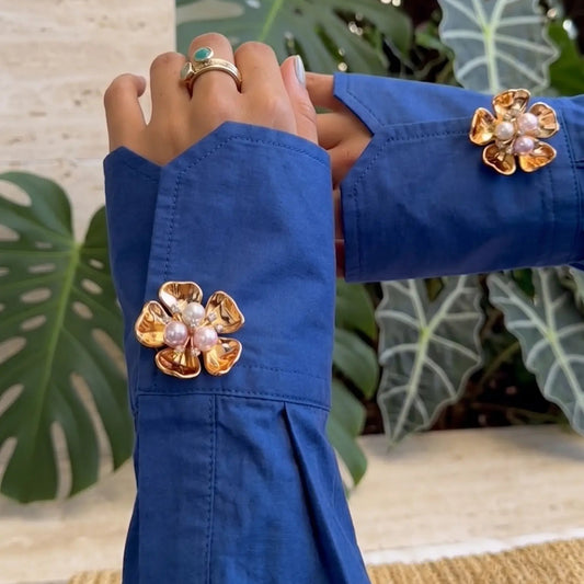 A vibrant lifestyle shot showing crossed arms wearing a bright blue shirt, elegantly styled with gold floral button covers featuring a multi-colored pearl cluster center. The lush tropical garden background perfectly highlights a chic resort wear aesthetic and everyday vacation luxury.