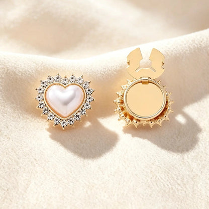 A delicate gold heart-shaped button cover featuring a luminous pearl center and a sparkling rhinestone halo. Displayed on a soft cream fabric, the right piece shows the open gold clasp mechanism, designed to instantly upgrade standard shirt buttons with a romantic, coquette aesthetic.