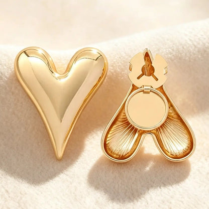 A sleek gold puffed heart button cover with a highly polished mirror finish, resting on soft cream fabric. The right piece displays the open clasp mechanism, showing how this minimalist faux cufflink easily attaches to standard shirt buttons.