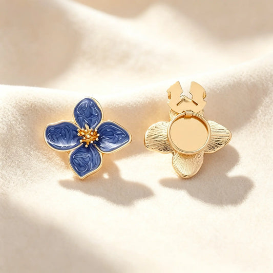 A close-up of a lapis blue marbled enamel four-petal floral button cover with a detailed textured gold stamen. Displayed on a soft cream fabric, the right piece shows the open gold clasp mechanism, demonstrating how this faux cufflink easily slides over standard buttons for a quiet luxury upgrade.