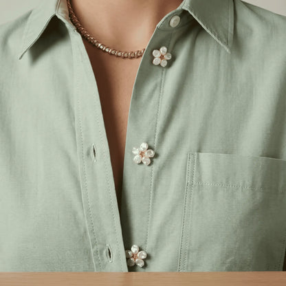 A lifestyle inspiration shot showing a woman wearing a crisp, light green cotton shirt, creatively styled with three delicately scaled pearl floral button covers down the front placket. These realistically proportioned faux cufflinks provide an effortless everyday quiet luxury upgrade without looking like heavy brooches.