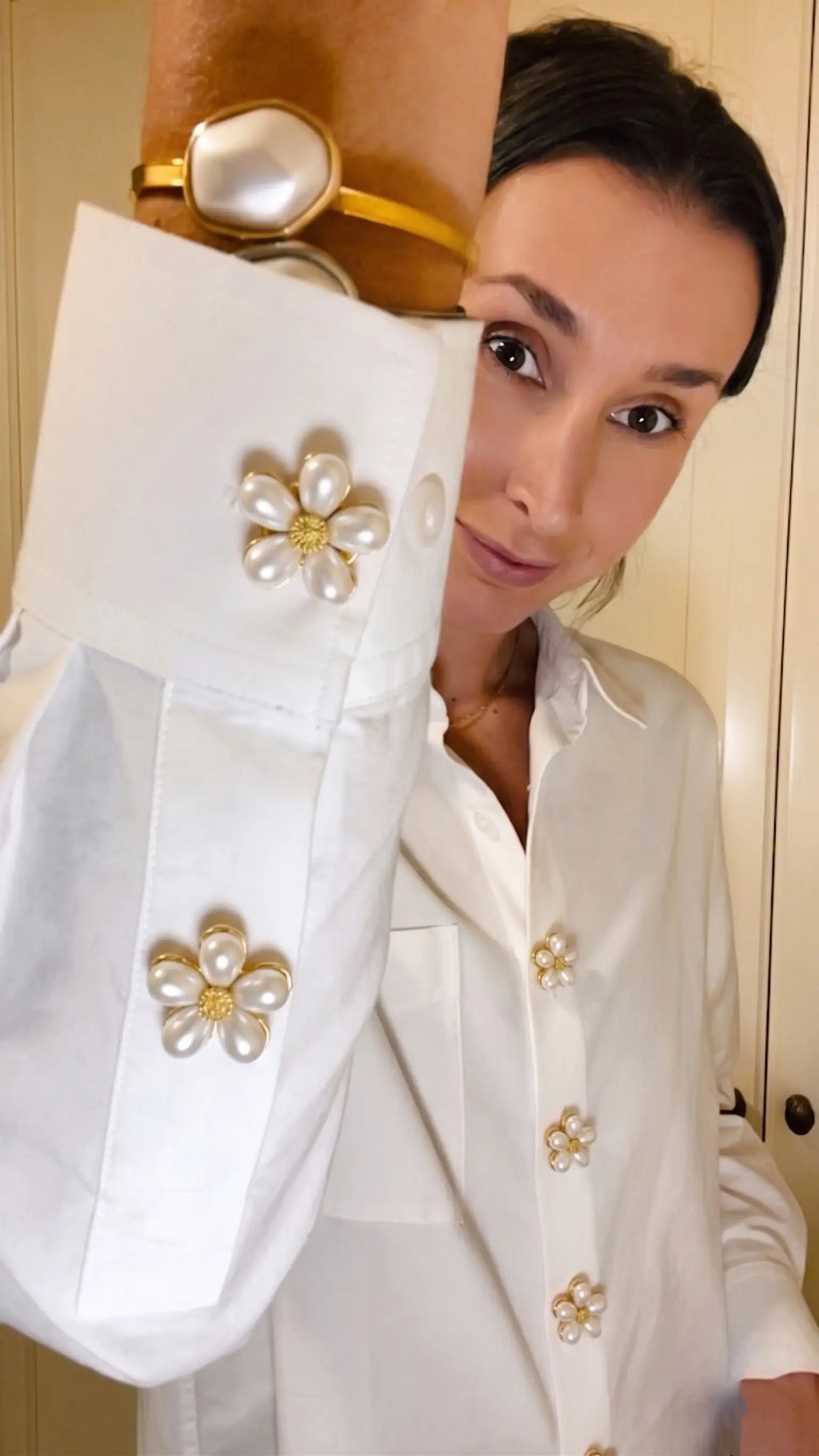 A woman wearing a classic white button-down shirt accessorized with multiple pearl floral button covers on the cuffs and front placket. A creative faux cufflink styling for a relaxed, everyday modern look.