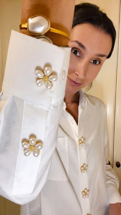 A woman wearing a classic white button-down shirt accessorized with multiple pearl floral button covers on the cuffs and front placket. A creative faux cufflink styling for a relaxed, everyday modern look.