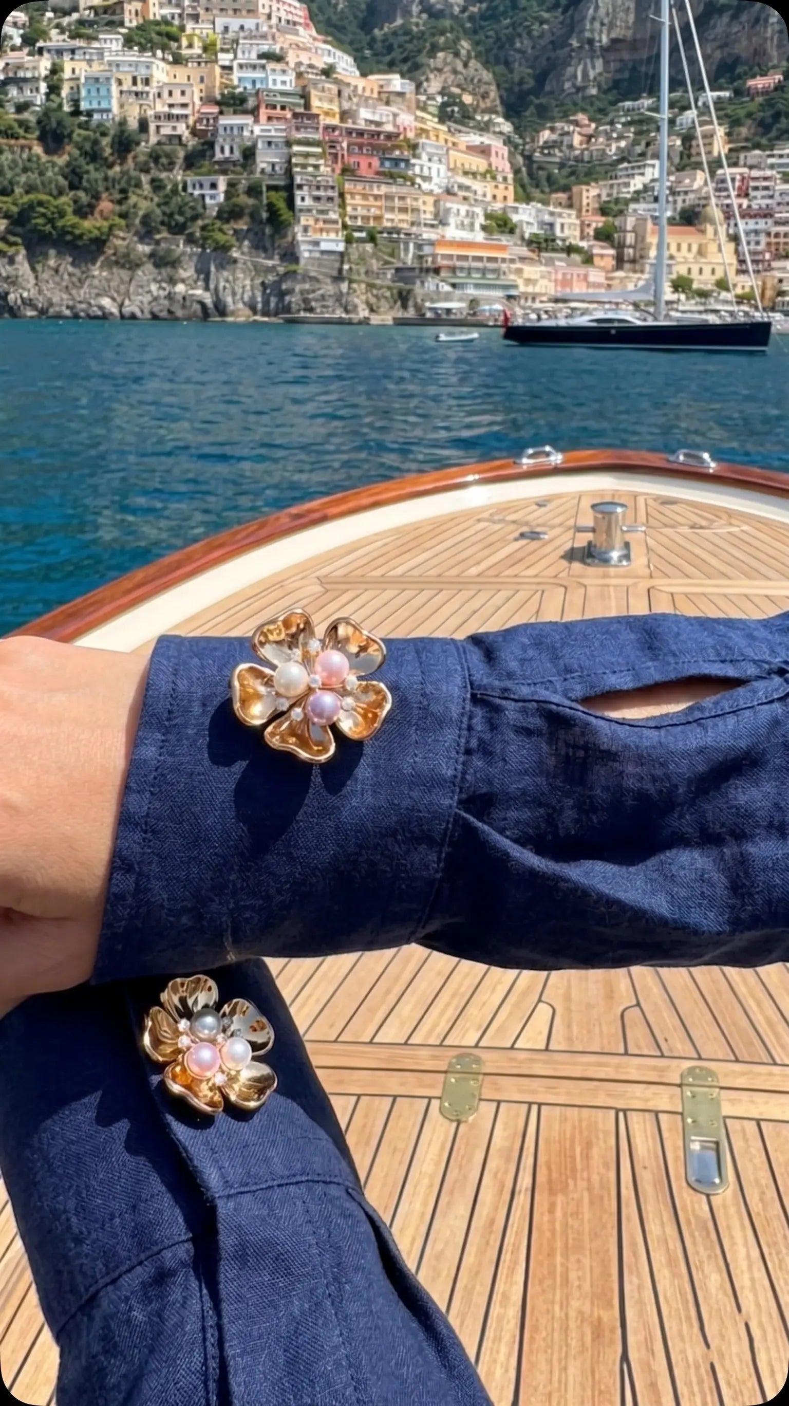 A breathtaking lifestyle photo of a woman wearing a navy blue linen shirt, elegantly styled with a gold floral button cover featuring a multi-colored pearl center. Set on a wooden yacht deck with a stunning Mediterranean coastal town in the background, showcasing the ultimate coastal luxury and holiday aesthetic.
