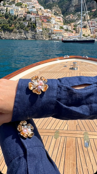 A breathtaking lifestyle photo of a woman wearing a navy blue linen shirt, elegantly styled with a gold floral button cover featuring a multi-colored pearl center. Set on a wooden yacht deck with a stunning Mediterranean coastal town in the background, showcasing the ultimate coastal luxury and holiday aesthetic.