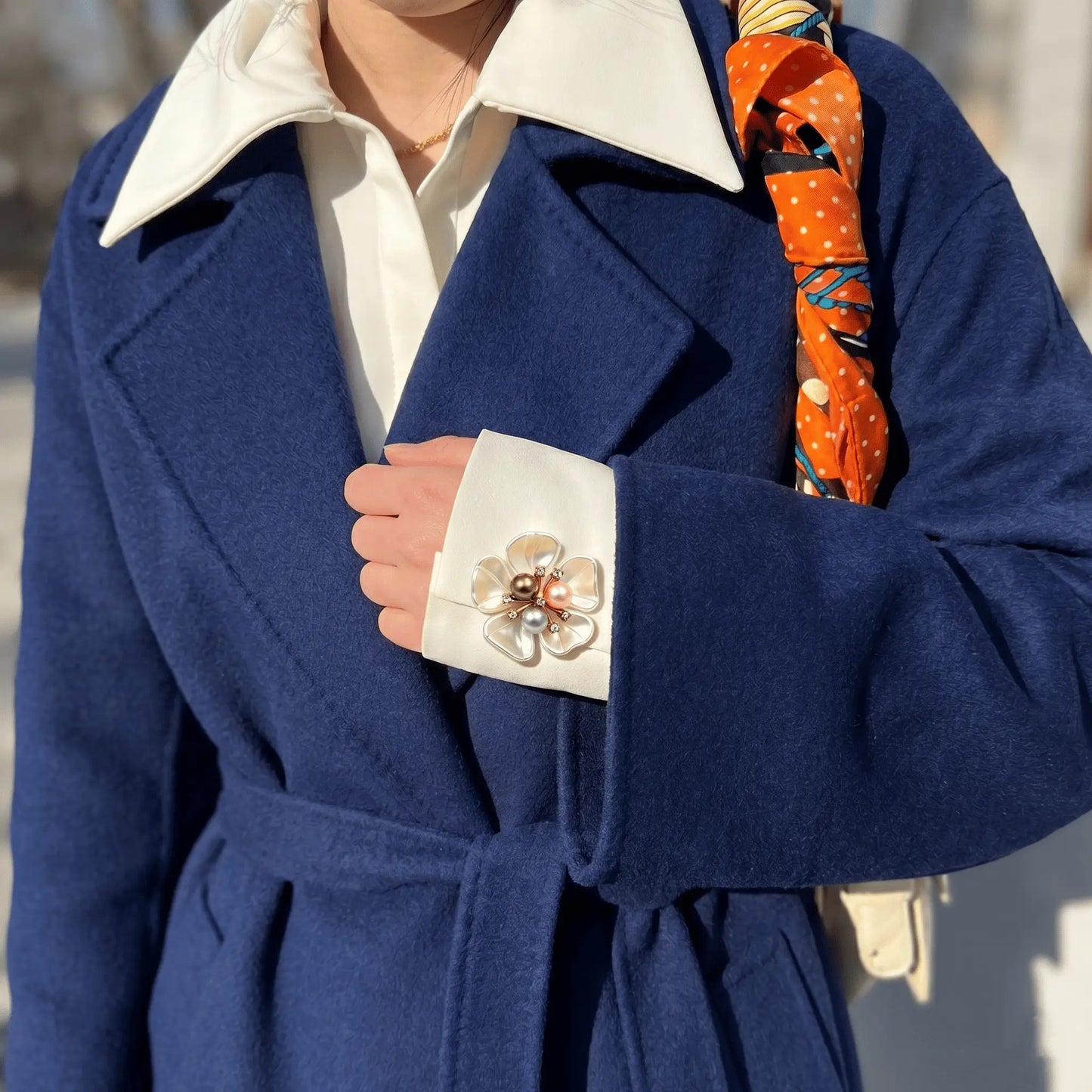 A chic winter street style shot of a woman wearing a navy blue coat with a colorful silk scarf. The crisp white shirt cuff is layered over the coat sleeve, beautifully accented by a pearlescent white floral button cover with mixed pearls. A perfect faux cufflink for elegant everyday layering.