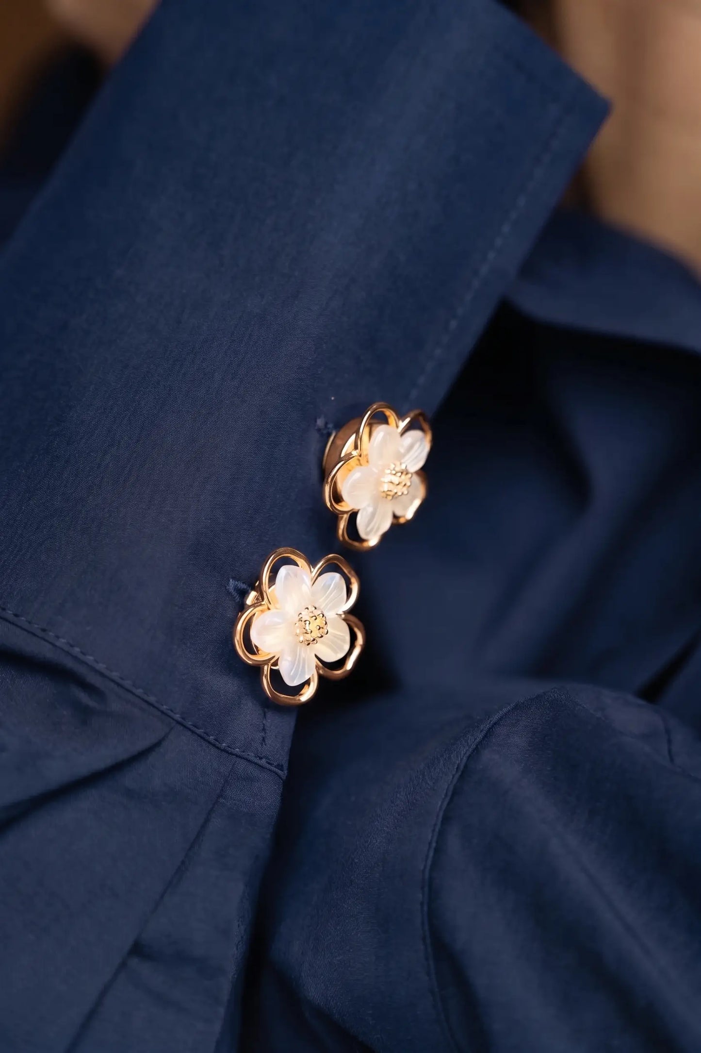 A detailed styling shot of a navy blue blouse featuring two pearlescent white floral button covers on the cuff. The luminous shell-like finish contrasts beautifully with the dark fabric, offering an easy, no-sew jewelry upgrade for everyday office wear.