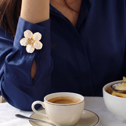 A chic everyday lifestyle shot of a woman enjoying a coffee break, wearing a luxurious navy blue silk shirt. Her cuff is elegantly styled with a realistically proportioned pearlescent white floral button cover. This delicately scaled faux cufflink seamlessly replaces a standard button, avoiding the heavy brooch look and adding a touch of quiet luxury to an effortless afternoon outfit.