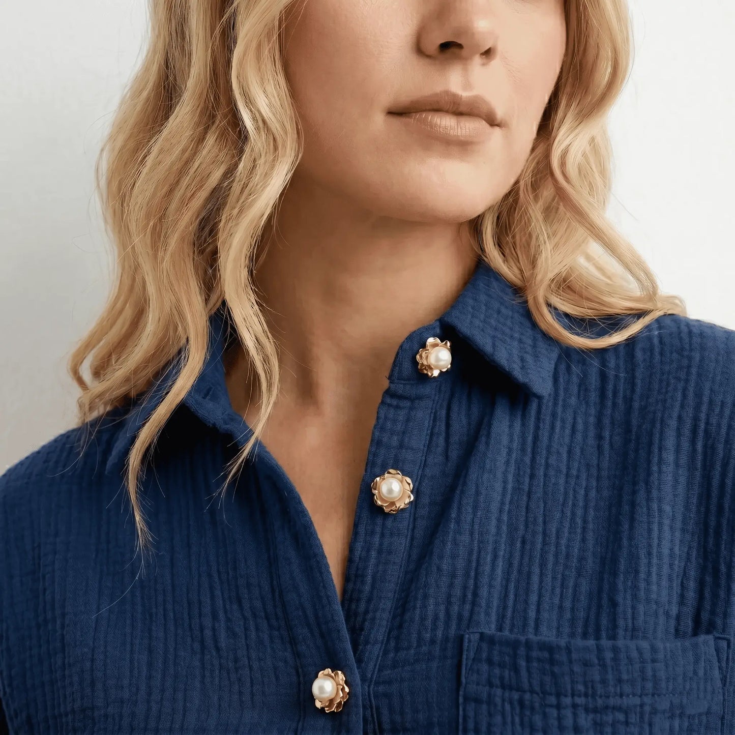 A sophisticated lifestyle shot of a woman styling three realistically scaled gold and pearl floral button covers down the front placket of a modern textured navy blue cotton shirt. These petite faux cufflinks offer an effortless everyday quiet luxury upgrade without looking like heavy brooches.