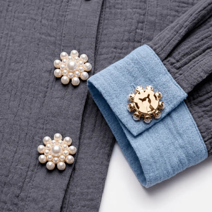 A creative flat lay styling shot featuring a modern textured grey cotton shirt with a light blue contrast cuff. Two realistically scaled pearl cluster sunflower button covers adorn the front placket, while a third is shown from the back, seamlessly securing the blue cuff with its gold clip. A versatile, light luxury faux cufflink that perfectly replaces standard buttons for everyday fashion.
