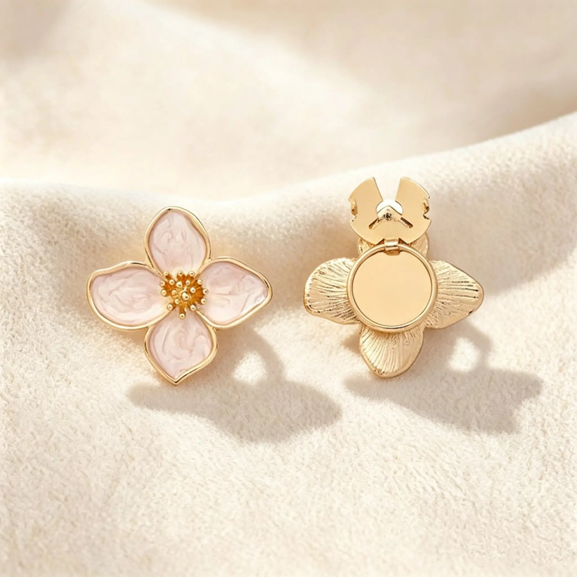 A delicate blush pink marbled enamel four-petal floral button cover featuring an intricate gold stamen center. Resting on soft cream fabric, the open gold clasp mechanism is clearly visible on the right, highlighting its effortless function as a realistic, no-sew faux cufflink.