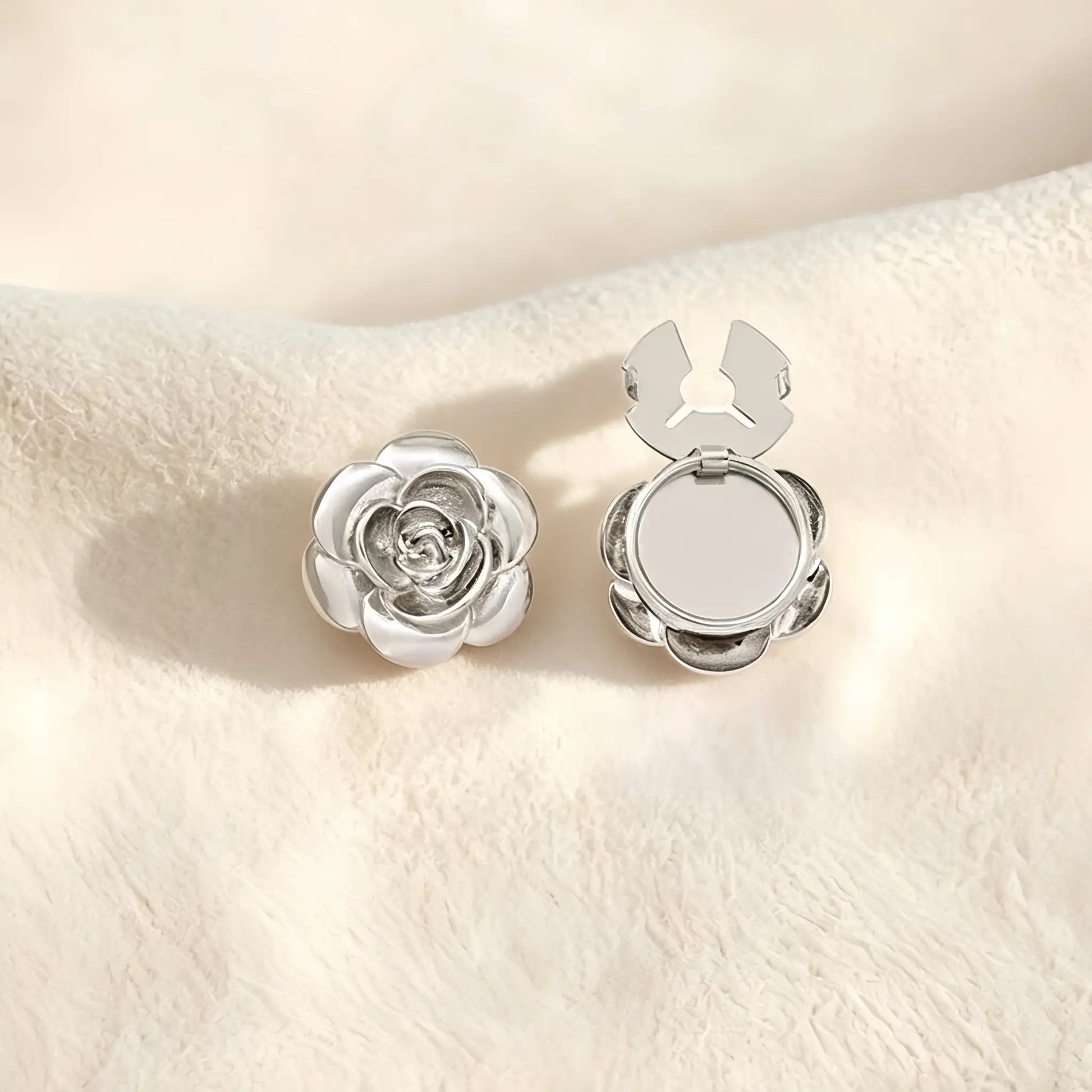 A sophisticated silver 3D rose button cover with delicate layered petals resting on a soft cream surface. The open silver clasp demonstrates the effortless functionality of this elegant faux cufflink accessory.