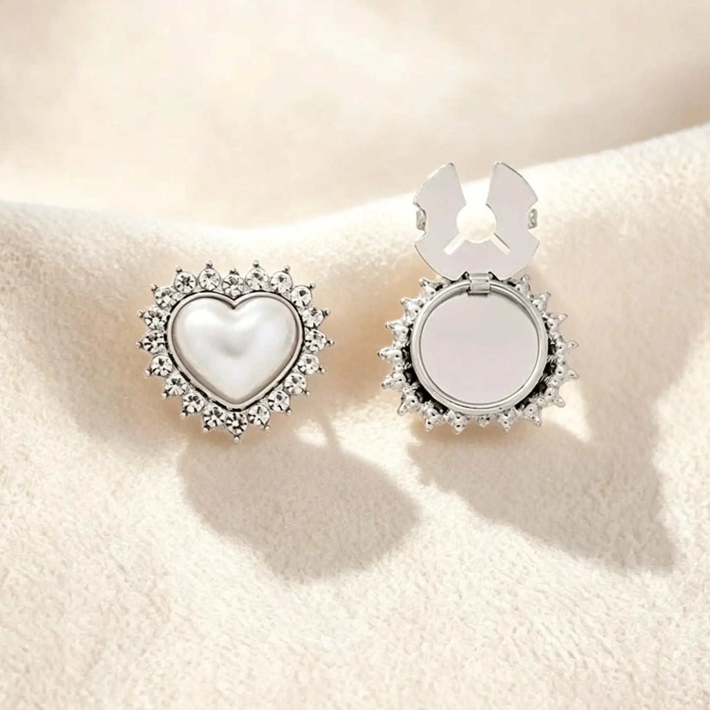 A sleek silver heart-shaped button cover showcasing a pearl core surrounded by a crystal rhinestone border. Resting on cream fabric, the right side displays the open silver attachment clip, offering a modern, cool-toned faux cufflink solution for everyday elegance.