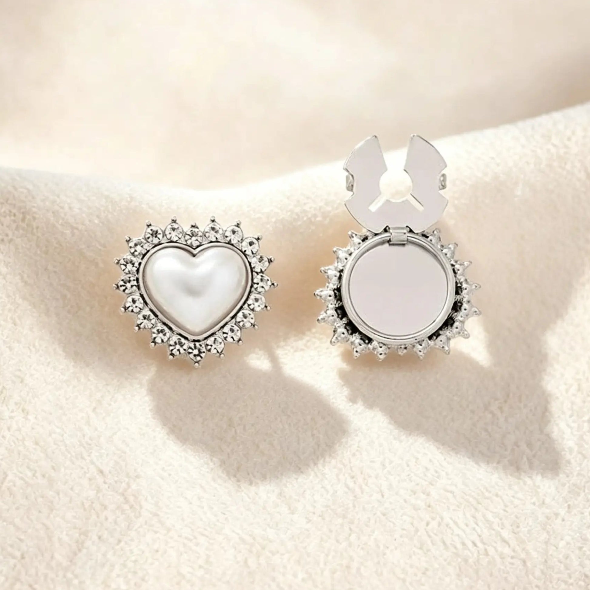 A sleek silver heart-shaped button cover showcasing a pearl core surrounded by a crystal rhinestone border. Resting on cream fabric, the right side displays the open silver attachment clip, offering a modern, cool-toned faux cufflink solution for everyday elegance.