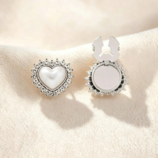A sleek silver heart-shaped button cover showcasing a pearl core surrounded by a crystal rhinestone border. Resting on cream fabric, the right side displays the open silver attachment clip, offering a modern, cool-toned faux cufflink solution for everyday elegance.