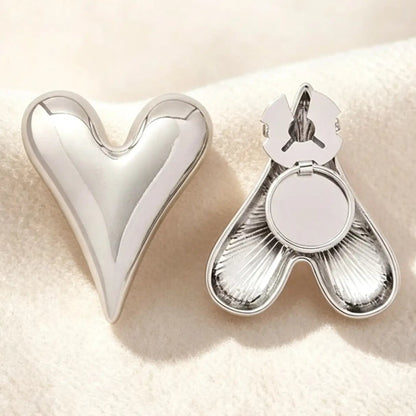 A contemporary silver puffed heart button cover displayed on textured cream fabric. The open backing on the right illustrates the clever no-sew clip design, perfect for transforming ordinary shirts into quiet luxury pieces.