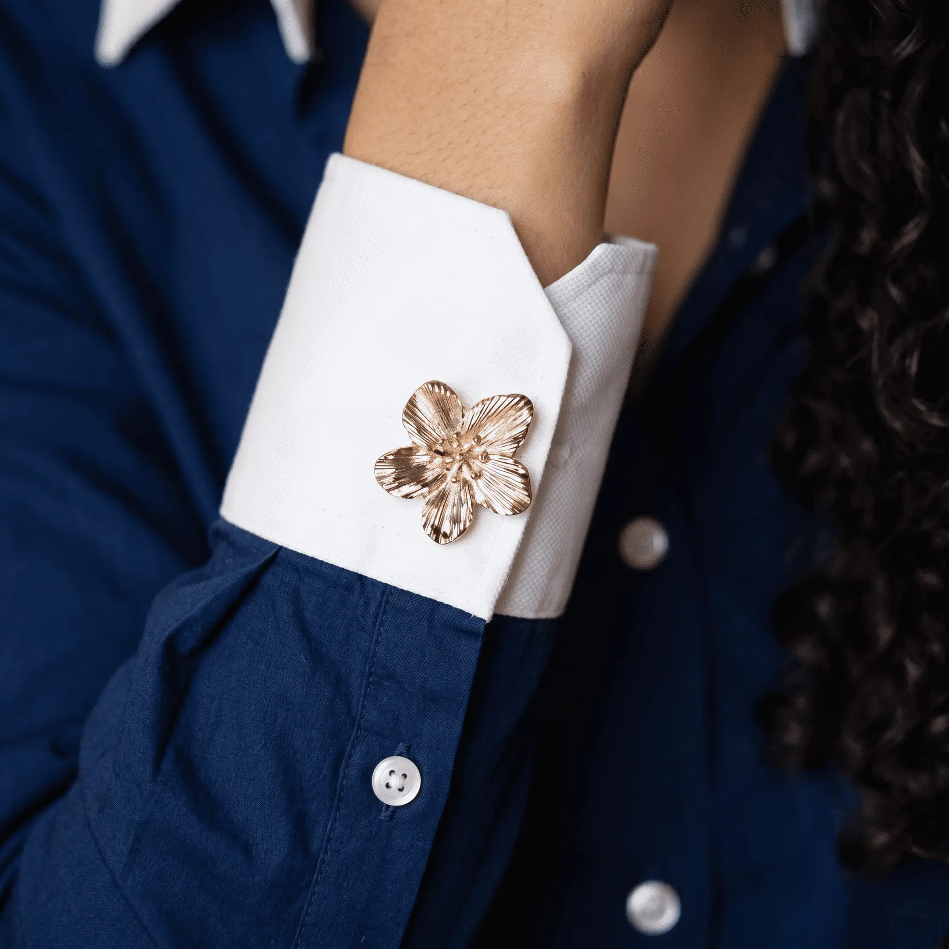A lifestyle shot of a woman wearing a modern navy blue cotton shirt with contrast white cuffs, elegantly styled with a realistically proportioned textured gold floral button cover. A perfect light luxury faux cufflink for everyday quiet luxury fashion.